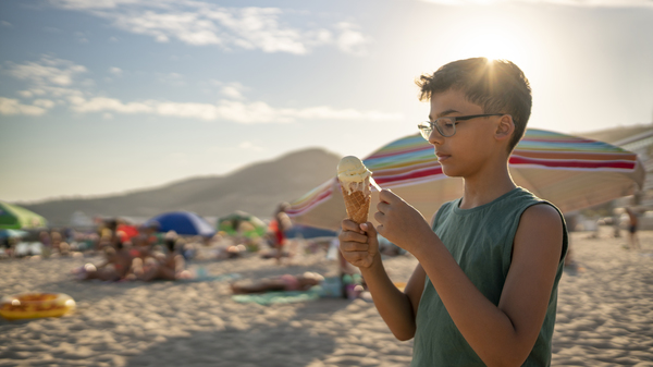 Boy eating ice cream in the Sun on a beach full of parasols and people.