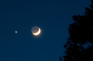 Crescent Moon with dark blue sky in the background. The unlit part of the Moon has a dim glow. Bright star shines to the left of the Moon.