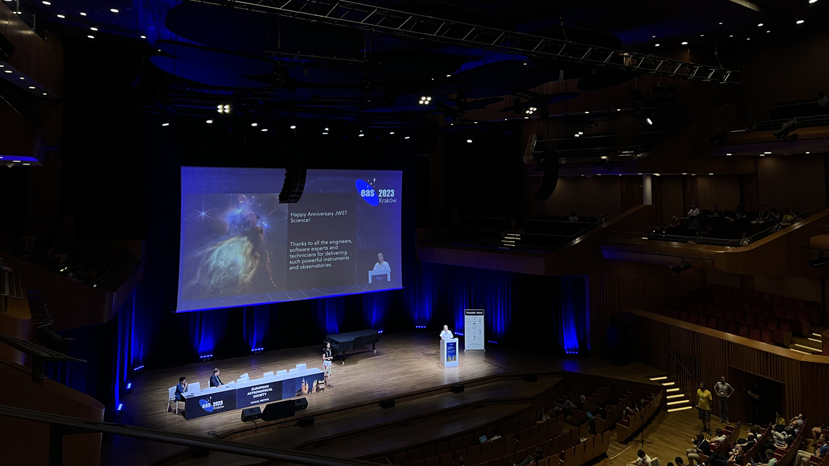 A dark conference room with high ceilings and a person presenting on stage.