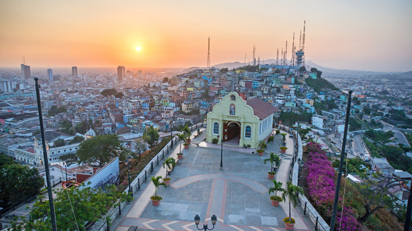 Panoramic sunset view of a hilltop church and cityscape, with colorful buildings, communication towers, and a large terrace overlooking the city.