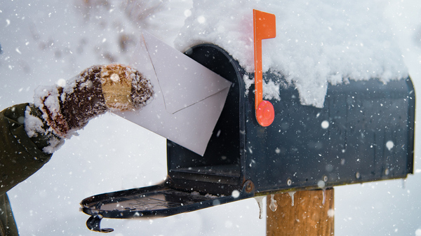 A gloved hand places a letter into a snow-covered mailbox, with snowflakes falling in a wintery scene.