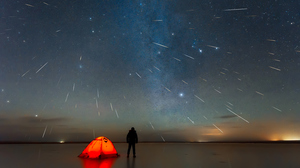 A person stands by an orange tent, watching meteors streak across a starry night sky under the Milky Way.