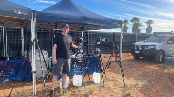 Man posing in front of several telescopes aimed at a blue sky.